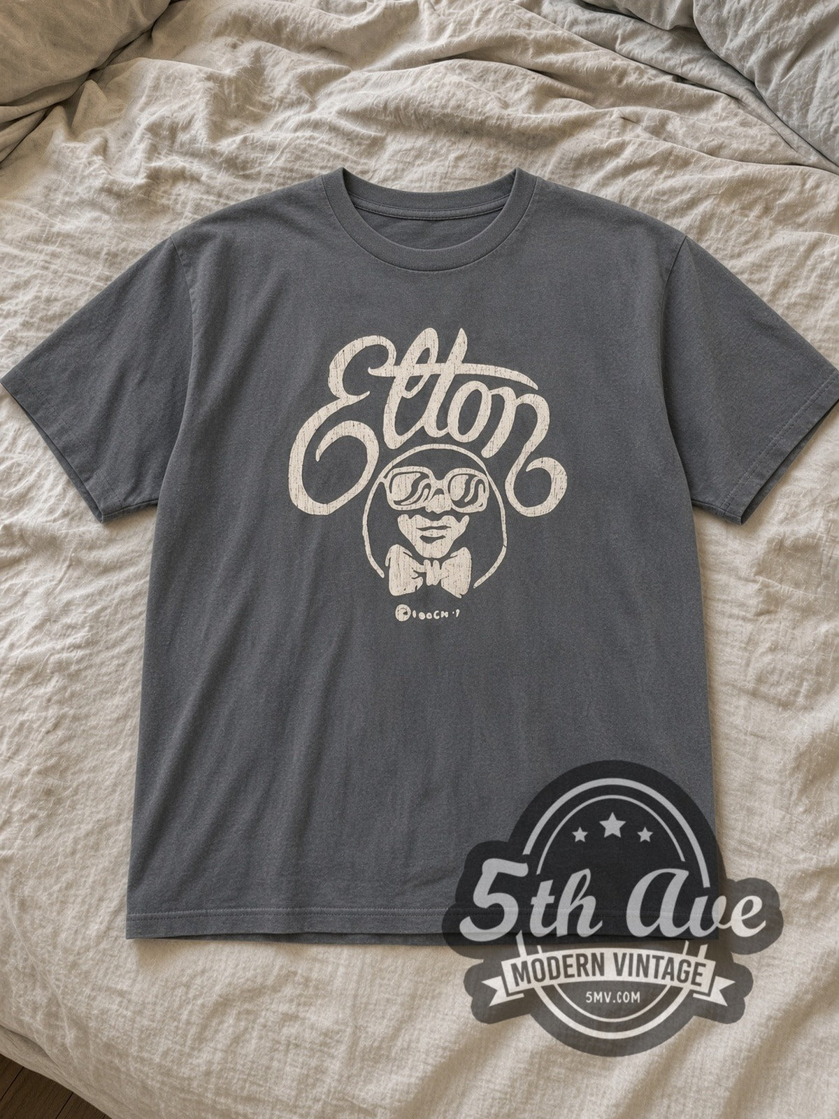 Charcoal Elton John Elton Script Cartoon Vintage Band T-Shirt laid flat on a linen bed